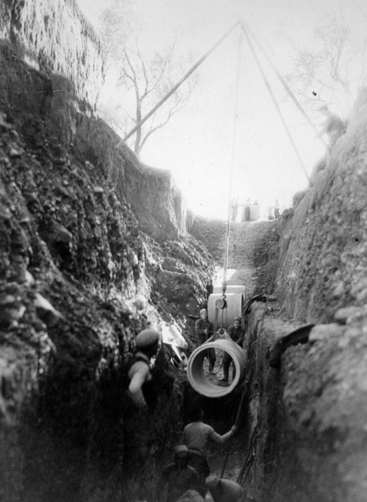A group of men working in the channel of a dry canal using a pulley system to install a drainage system.
