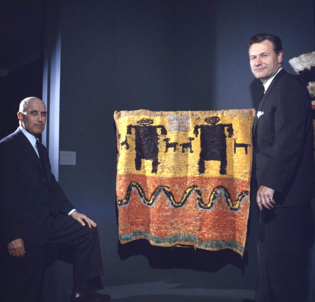 Robert Goldwater (left) and Nelson Rockefeller (right) posing in front of a tapestry at the Museum of Primitive Art