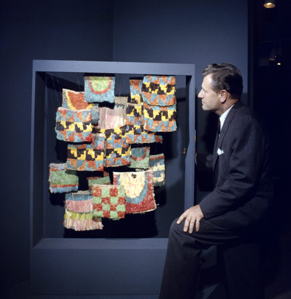 Nelson Rockefeller in front of a display of colorful hanging art at the Museum of Primitive Art.