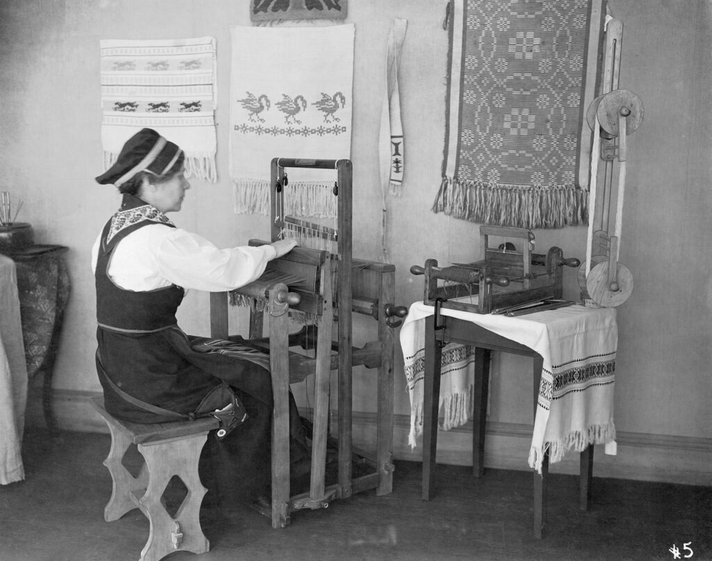 A woman in traditional eastern European dress sits and works at a loom, surrounded by examples of traditional needlework.