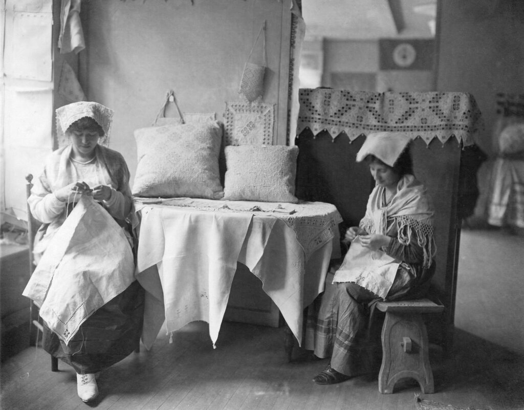 Two women sit at a table together each working individually on a piece of needlework.