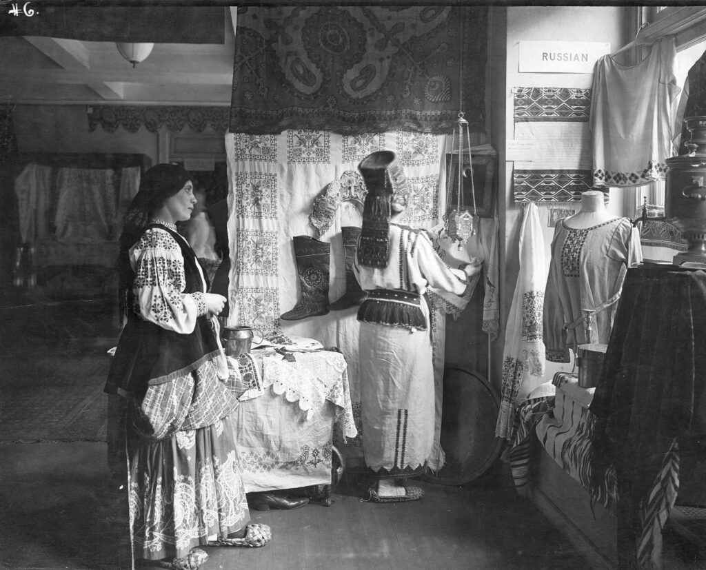 Two women in traditionally embroidered Eastern European dress tend to an exhibit of embroidered decor and garments.