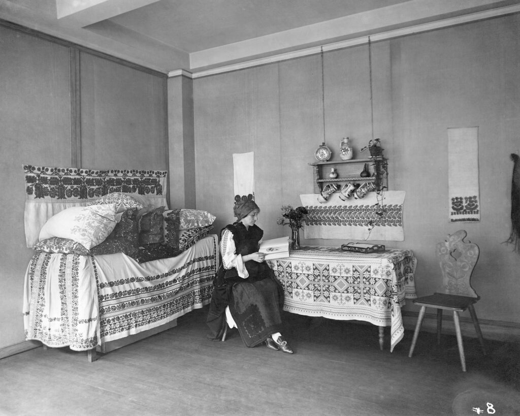 A woman in traditional dress sits reading a book surrounded by a display of embroidered home decor, such as pillows, tablecloths, hand towels, and wall tapestries.